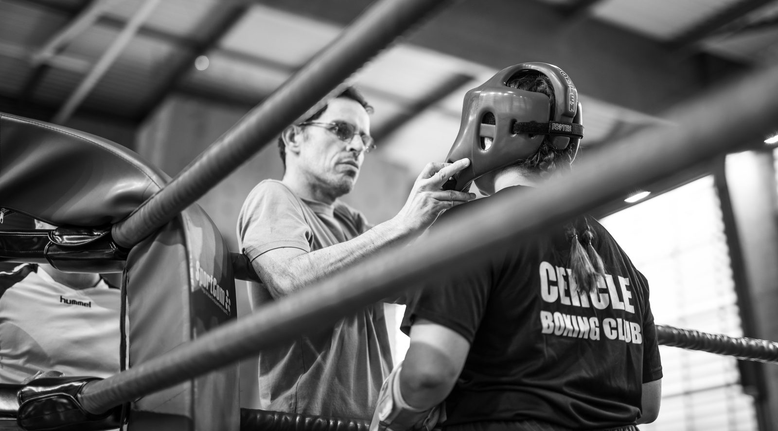 Un coach ajuste le casque d'un tireur, vu à travers les cordes du ring